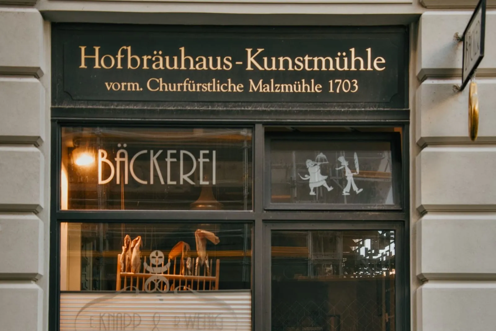 Die Front einer Bäckerei mit vergoldeter Beschriftung über dem Schaufenster, dahinter Gebäck auf einem Holzgestell