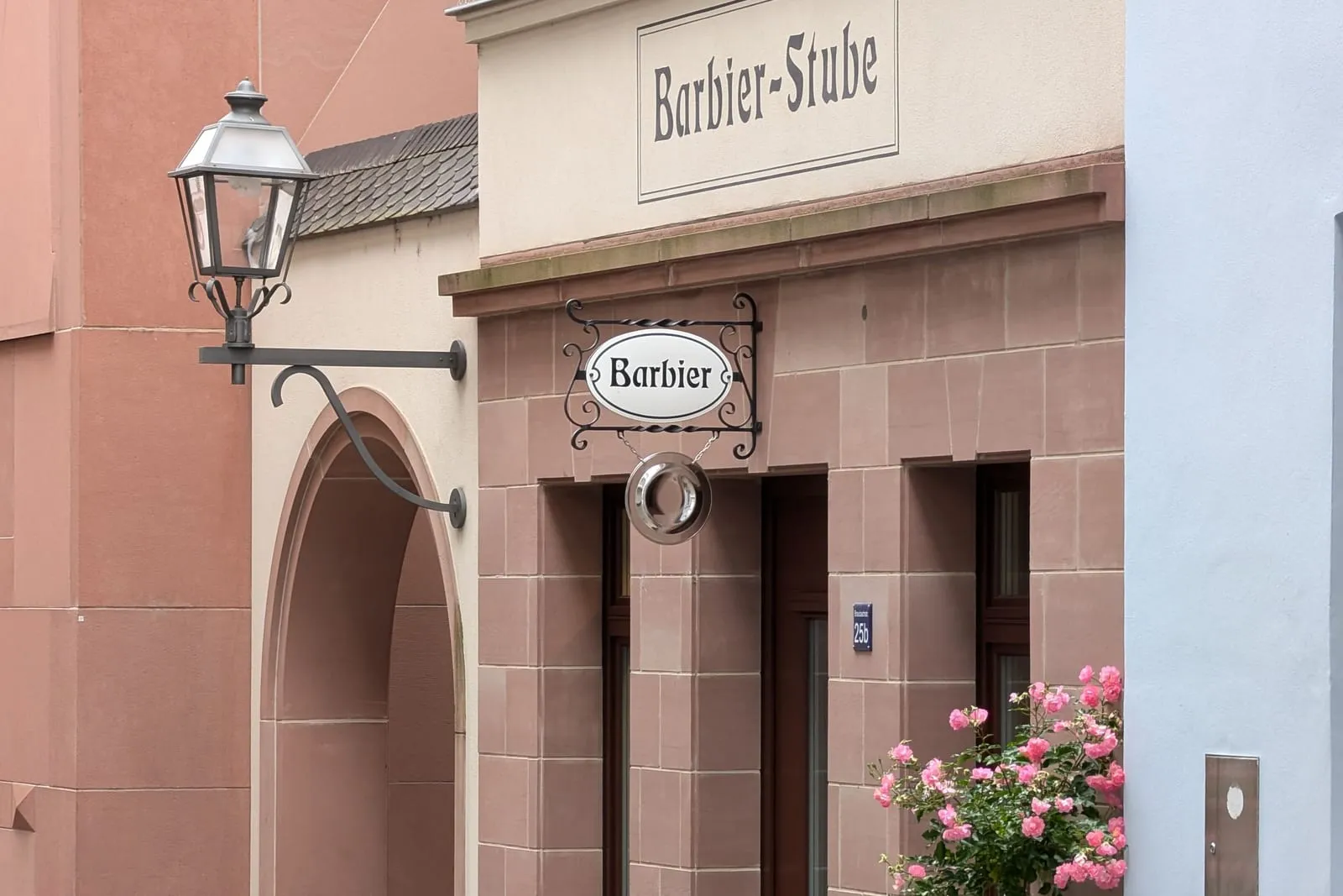 Ein schmiedeeisernes Auslegerschild mit der Aufschrift Barbier an einer Sandsteinfassade, daneben eine Straßenlaterne