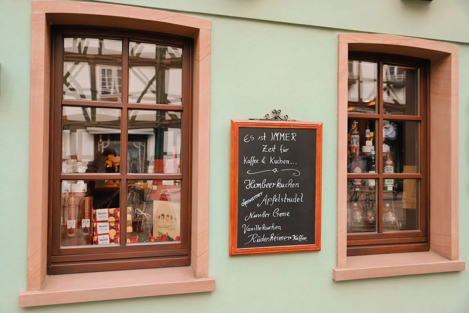Eine handbeschriebene Kreidetafel mit dem Kuchenangebot des Tages, zwischen zwei Schaufenstern einer grün gestrichenen Hausfassade